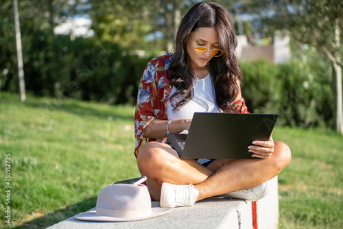 Woman with sunglasses using a laptop computer while sitting on a bench in a park on a sunny day. © Luis