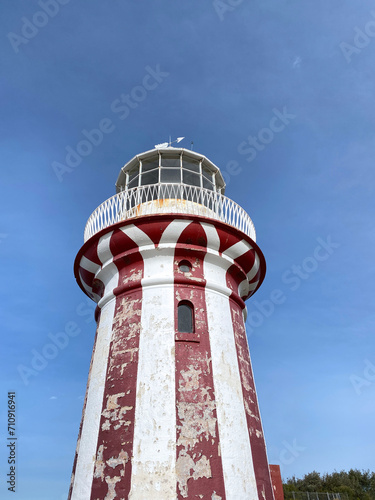 Red lighthouse with white stripes on blue sky. Coast lighthouse at the top of a hill. Harbor in the island.