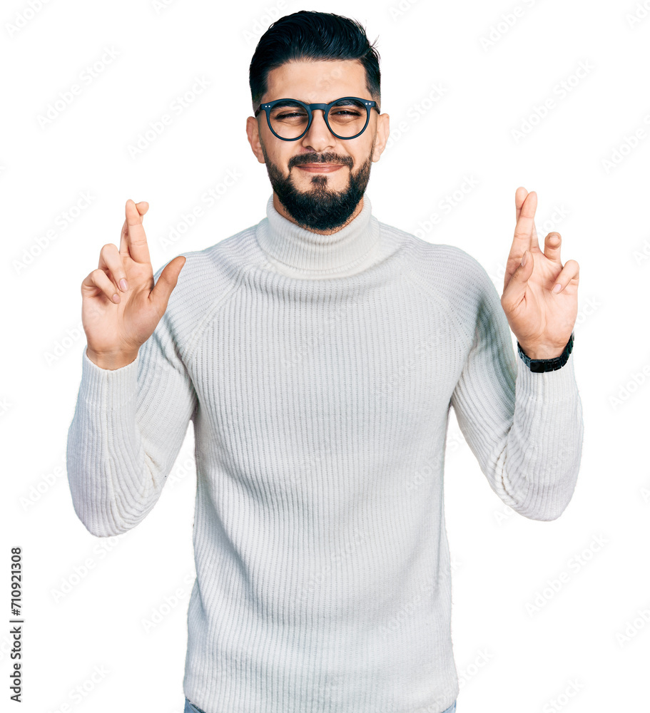 Fototapeta premium Young arab man with beard wearing elegant turtleneck sweater and glasses gesturing finger crossed smiling with hope and eyes closed. luck and superstitious concept.