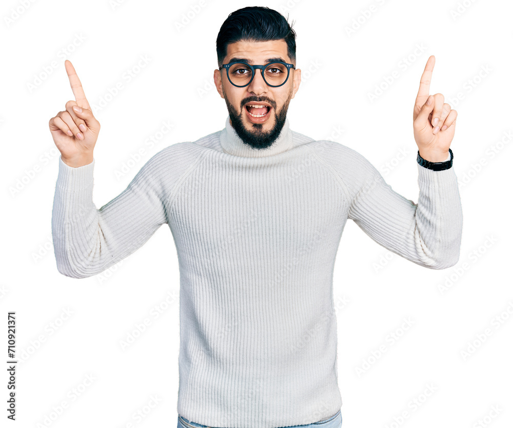 Young arab man with beard wearing elegant turtleneck sweater and glasses smiling amazed and surprised and pointing up with fingers and raised arms.