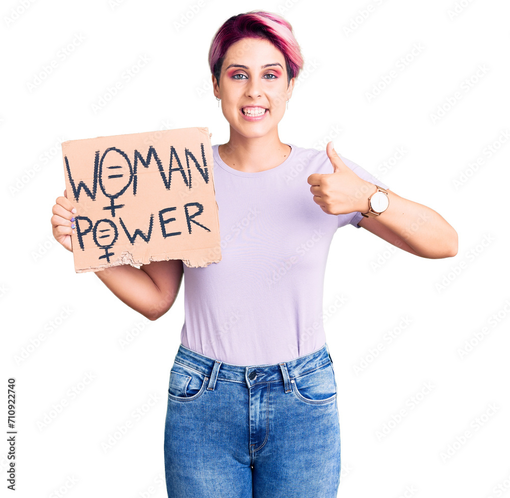 Young beautiful woman with pink hair holding woman power banner smiling happy and positive, thumb up doing excellent and approval sign Young beautiful woman with pink hair holding woman power banner smiling happy and positive, thumb up doing excellent and approval sign
