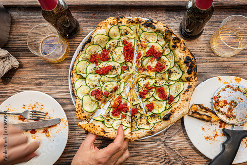 Male hand taking a slice of freshly baked Neapolitan zucchini pizza on a rustic wooden table in a traditional Pizzeria. photo