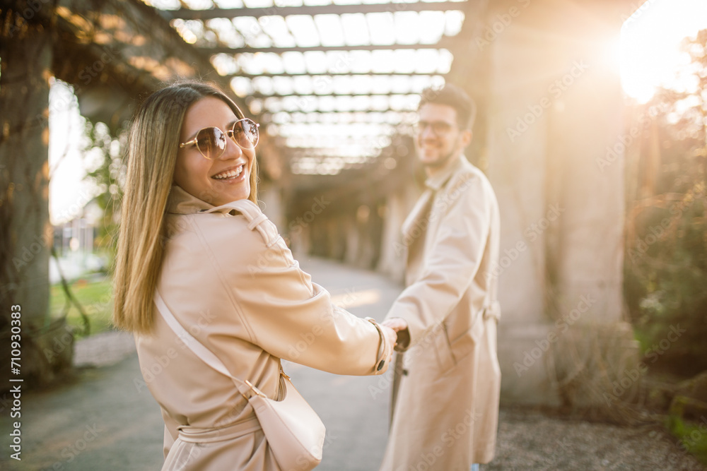 Fototapeta premium A stylish couple walks through the park in beige trench coats. A woman and a man spend time together in the spring at sunset. Concept of beauty, love and happiness.