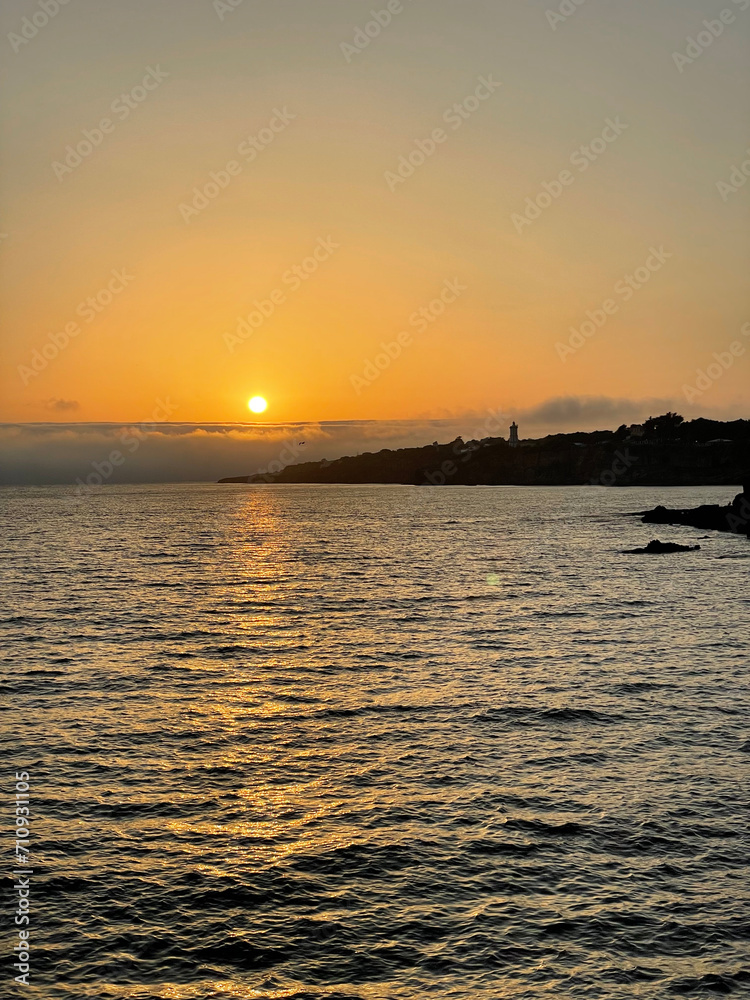 Toma vertical de una puesta de sol en el mar con la silueta de un faro en el horizonte, en tonos plateados y naranjas