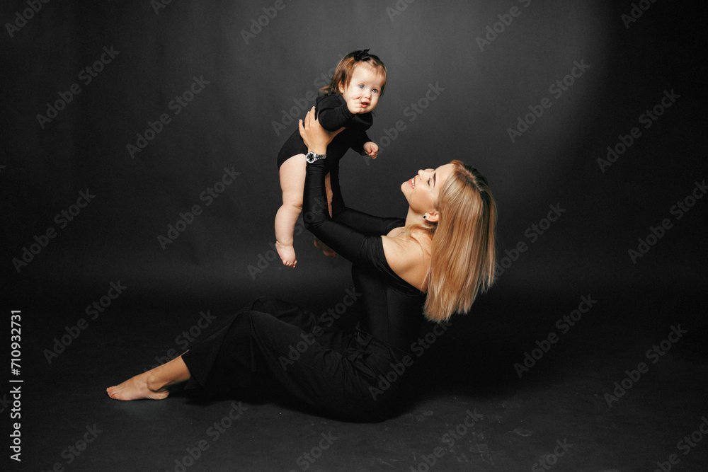 Portrait of a young beautiful mother holding her baby daughter in her arms, hugging and smiling while sitting in a studio on a black background