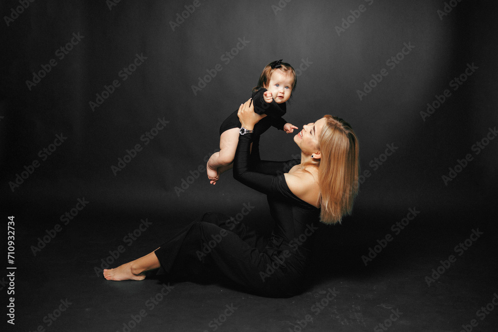 Close-up portrait of a young smiling mother holding and hugging her toddler daughter on a black background. Happy Relationship of mother and small child. Mother is playing with the baby girl Close-up portrait of a young smiling mother holding and hugging her toddler daughter on a black background. Happy Relationship of mother and small child. Mother is playing with the baby girl