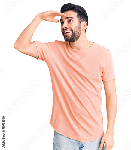 Young handsome man with beard wearing casual t-shirt very happy and smiling looking far away with hand over head. searching concept.