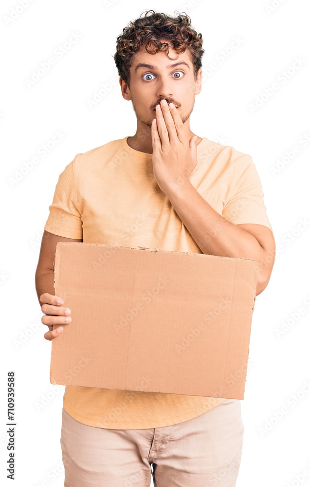 Young handsome man with curly hair holding blank cardboard empty banner covering mouth with hand, shocked and afraid for mistake. surprised expression Young handsome man with curly hair holding blank cardboard empty banner covering mouth with hand, shocked and afraid for mistake. surprised expression