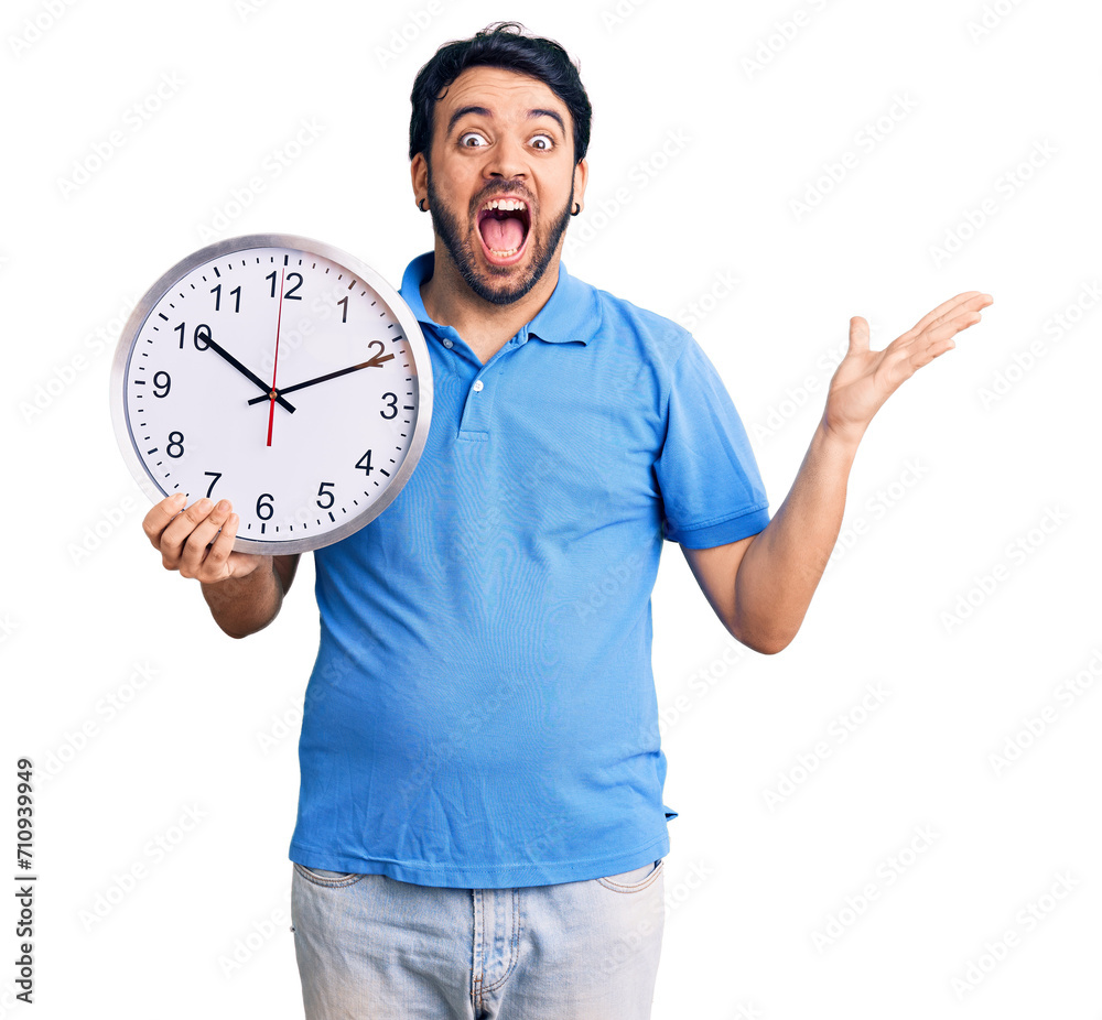 Young hispanic man holding big clock celebrating victory with happy smile and winner expression with raised hands Young hispanic man holding big clock celebrating victory with happy smile and winner expression with raised hands