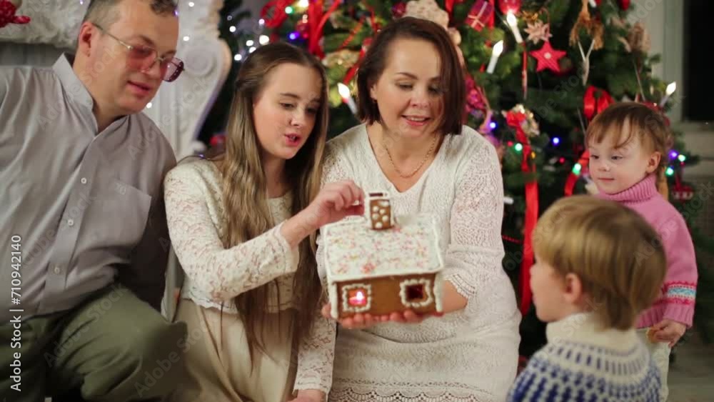 Family of five looks on gingerbread house near Christmas tree Family of five looks on gingerbread house near Christmas tree
