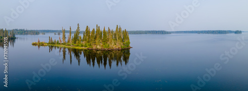 An aerial panoramic view of a large calm lake with a small island that is covered in a forest of spruce and pine trees. The calm water reflects the trees and the blue sky. 