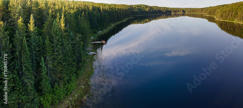 Aerial panoramic view of a small calm northern lake that is surrounded by spruce trees. The morning light highlights the tops of the trees. The peaceful calm water reflects a blue sky with scattered w