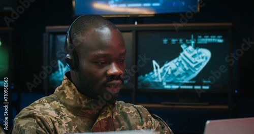 Close up of African American young man in headset sitting at laptop computer and talking via videochat in monitoring room in army. Soldier having chat via webcam in military center. photo