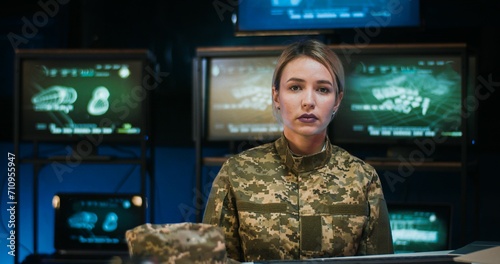 Portrait of pretty Caucasian female soldier sitting at desk in monitoring room, taking on cap and looking at camera. Attractive woman in militarian uniform working in troops. Girl in army concept. photo
