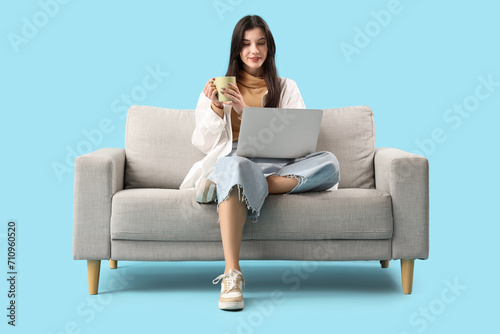Beautiful young woman with cup of tea and laptop on sofa against blue background