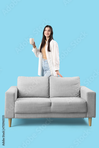 Beautiful young woman with cup of tea and sofa on blue background