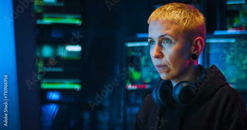 Portrait of Caucasian woman hacker with blond short hair sitting in dark room with monitors at desk in front of computer. Turning face to camera and taking on hood. Close up of female hacking system.