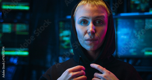 Close up of Caucasian woman with short blonde hair looking straight to camera in dark IT office and taking on black hood. At night. Portrait of female anonymous hacker concept. photo