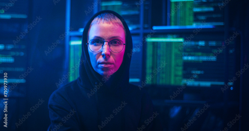 Caucasin female hacker in dark hood and glasses turning to camera on spinning chair and looking straight at night in room with monitors and data. Portrait of woman sittng in cyber police offce. Caucasin female hacker in dark hood and glasses turning to camera on spinning chair and looking straight at night in room with monitors and data. Portrait of woman sittng in cyber police offce.