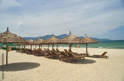 Fototapeta Naklejka Na Ścianę i Meble -  Straw sunshades and sunbeds on the empty pebble beach with sea in the background. Deserted beach with rattan sun loungers and umbrellas