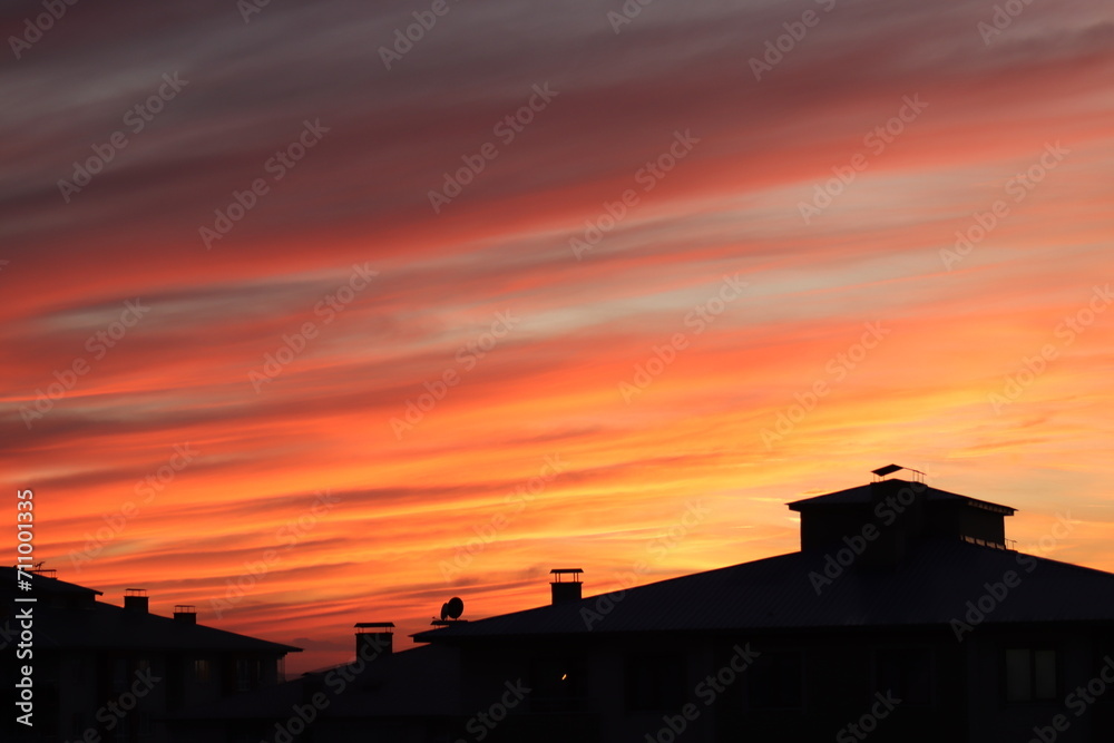 A dramatic sunset with radiant hues of orange and red streaking across the sky, silhouetting the rooftops of urban buildings.
