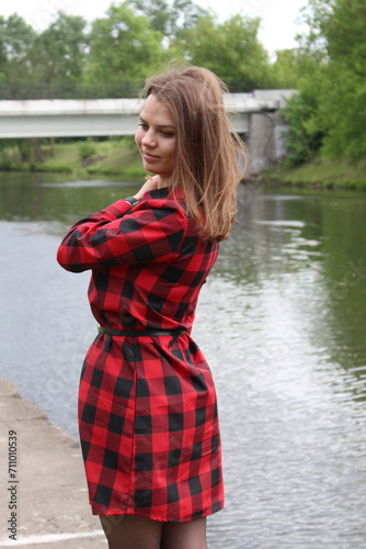 A young girl in a red and black dress walks against the background of a pond in the city in summer