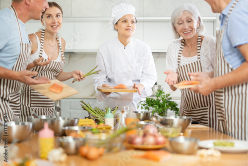 Friendly skilled young Asian female chef giving culinary classes to group of men and women of different ages  teaching to cook salmon deliciously