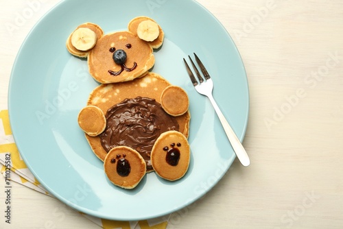 Creative serving for kids. Plate with cute bear made of pancakes, blueberries, bananas and chocolate paste on light wooden table, top view. Space for text