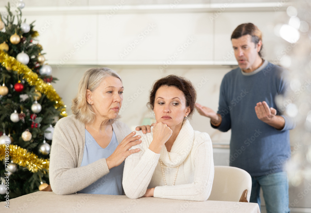 Husband standing near christmas tree sorts things out with his thinking wife in bright kitchen, and elderly woman calms down sad woman