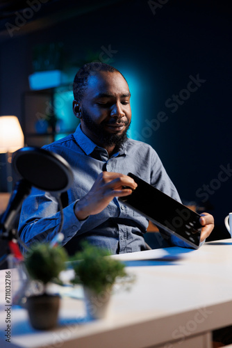 Tech engineer presenting tablet in entry level low budget price range, filming from dimly lit home studio. Viral tech content creator reviewing digital device in front of online audience