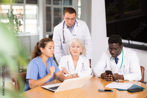 Three physicians sitting and young nurse standing by them looking at the notebook interestedly