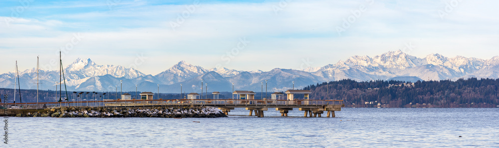 A fishing pier extends out into the ocean with a backdrop of mountains and a cloudy sky