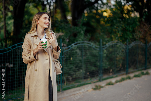 Woman holding a cup of coffee standing on a city street next to a park.