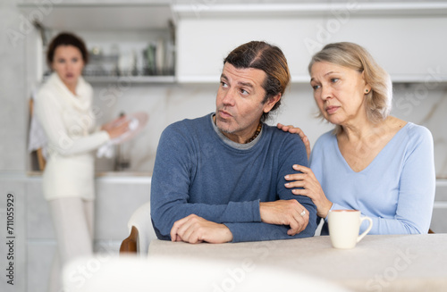 Elderly woman sits next to her adult son, pats hand and comfort him during family quarrel with his wife. Angry woman screams at her husband