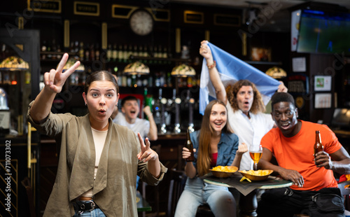 Happy young girl emotionally gesturing while watching football match in sports bar, cheering for favorite team of Argentina and celebrating victory