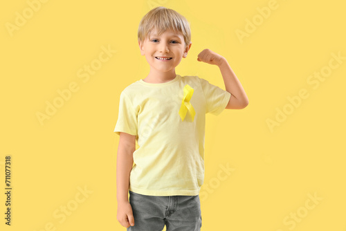 Cute little boy with yellow ribbon showing muscles on color background. Childhood cancer awareness concept