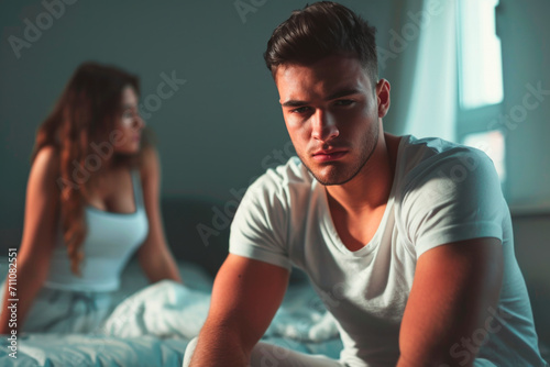 guy sitting on the edge of his bed pensive, looking down, in the background a woman looking through the window 