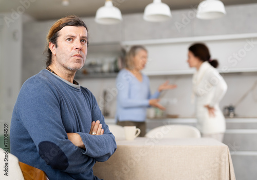 Frustrated man sitting in kitchen during quarrel between his wife and elderly mother. Complicated family relationships, daughter-in-law and mother-in-law yell at each other © JackF