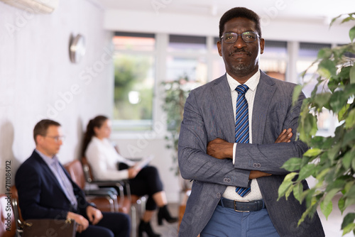 Friendly secretary African-American man stands at entrance to office reception with folder and list of guests and expects invited visitors. Male office hostess willing to help photo