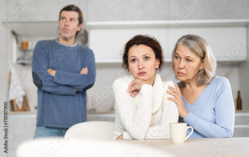 Upset woman sits near kitchen table with mother and listens to discontent and indignation of older brother. Senior woman supports comforting and calms daughter during quarrel with relative