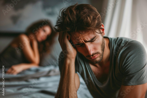 guy sitting on the edge of his bed sad, looking down, in the background a woman looking at him 
