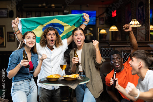 Enthusiastic multiracial group of young adult sports fans celebrating victory of favorite team with beer in bar, waving national flag of Brazil