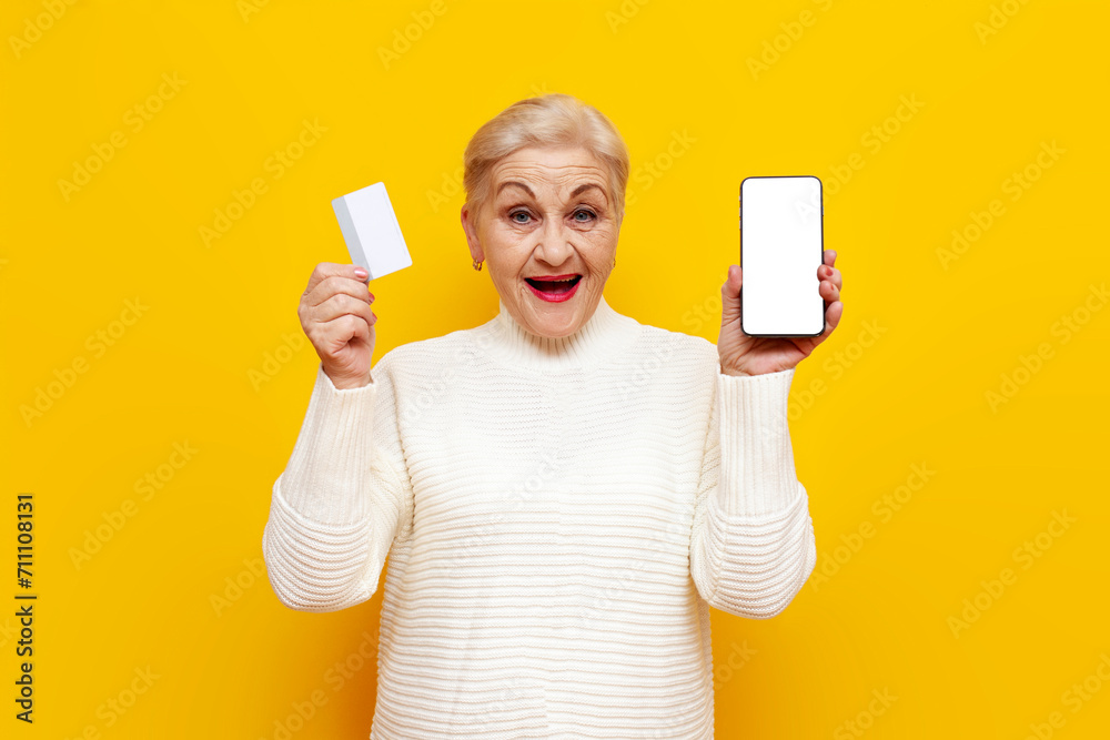old grandmother in a white sweater shows a credit card and a blank smartphone screen on a yellow isolated background, elderly woman pensioner buys online - obrazy, fototapety, plakaty 