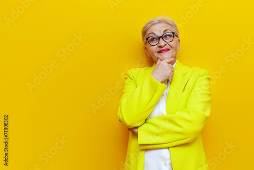 old thoughtful grandmother in business clothes blazer plans and thinks on a yellow isolated background, elderly businesswoman in a suit imagines and imagines looking away