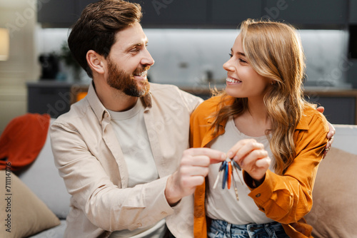 Happy young family holding keys from new home looking each other. Attractive smiling man and beautiful woman hugging buying new property sitting together on comfortable sofa. Moving concept