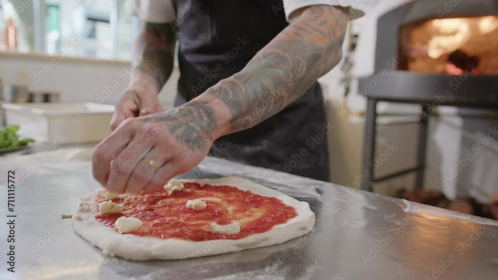 Hands of chef in apron standing behind the table in commercial kitchen and adding cheese on top of pizza base with tomato sauce. Close-up shot