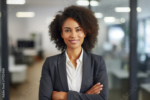 Business woman. Portrait of a african american, beautiful, young and happy woman in a suit standing in a modern office. Smiling female manager looking at the camera in a workplace