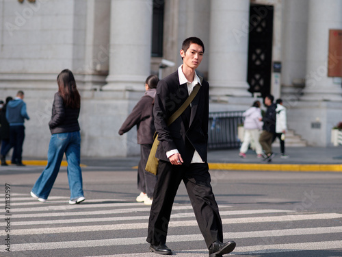 Portrait of handsome Chinese young man with black short hair wearing black blazer walking on street with modern city building background in sunny winter day, male fashion, cool Asian young man.