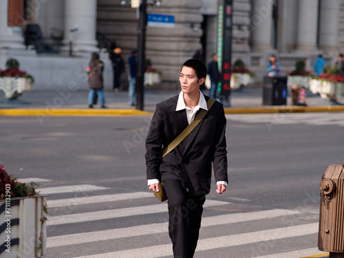 Portrait of handsome Chinese young man with black short hair wearing black blazer walking on street with modern city building background in sunny winter day, male fashion, cool Asian young man.
