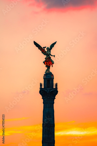 The Angel of Independence statue placed on Promenade of the Reform in Mexico city between tall buildings against colorful orange sunset sky in evening